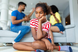 Black girl looking sad on the floor. Parents fighting in the background. Child suffering from quarrels between parents in the family at home. Domestic violence and Family conflict concept. Sadness little girl against blured of mother fighting father with quarrel at home.